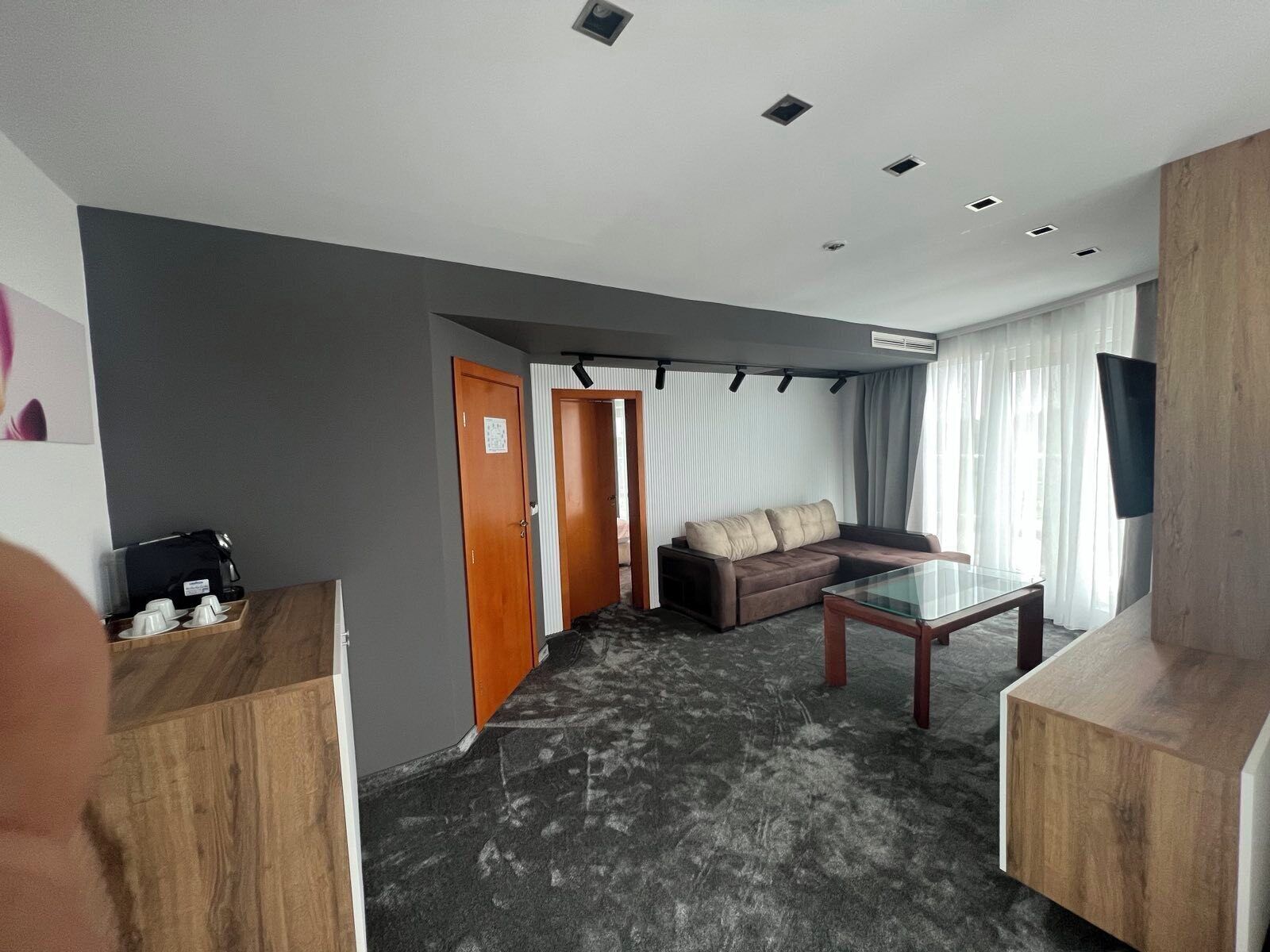 A modern hotel room with a sofa, a glass coffee table, a TV mounted on the wall, and a door leading to another room.