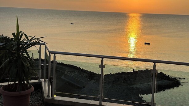 Une vue sur le coucher de soleil depuis un balcon doté d’une balustrade, donnant sur une mer calme et une côte rocheuse.