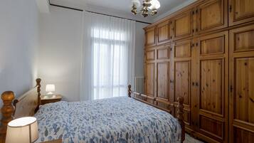 Una camera da letto con un'ampia armadiatura in legno, un letto con copripiumino azzurro decorato, un comodino con una lampada e una finestra con tende leggere.