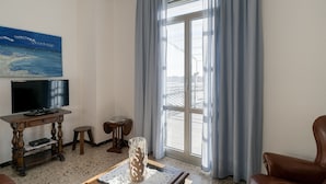 A living room with a television, a wooden table, a glass vase, a chair, and a large window with curtains. - Agriturismo Le Girandole (Diano Marina)