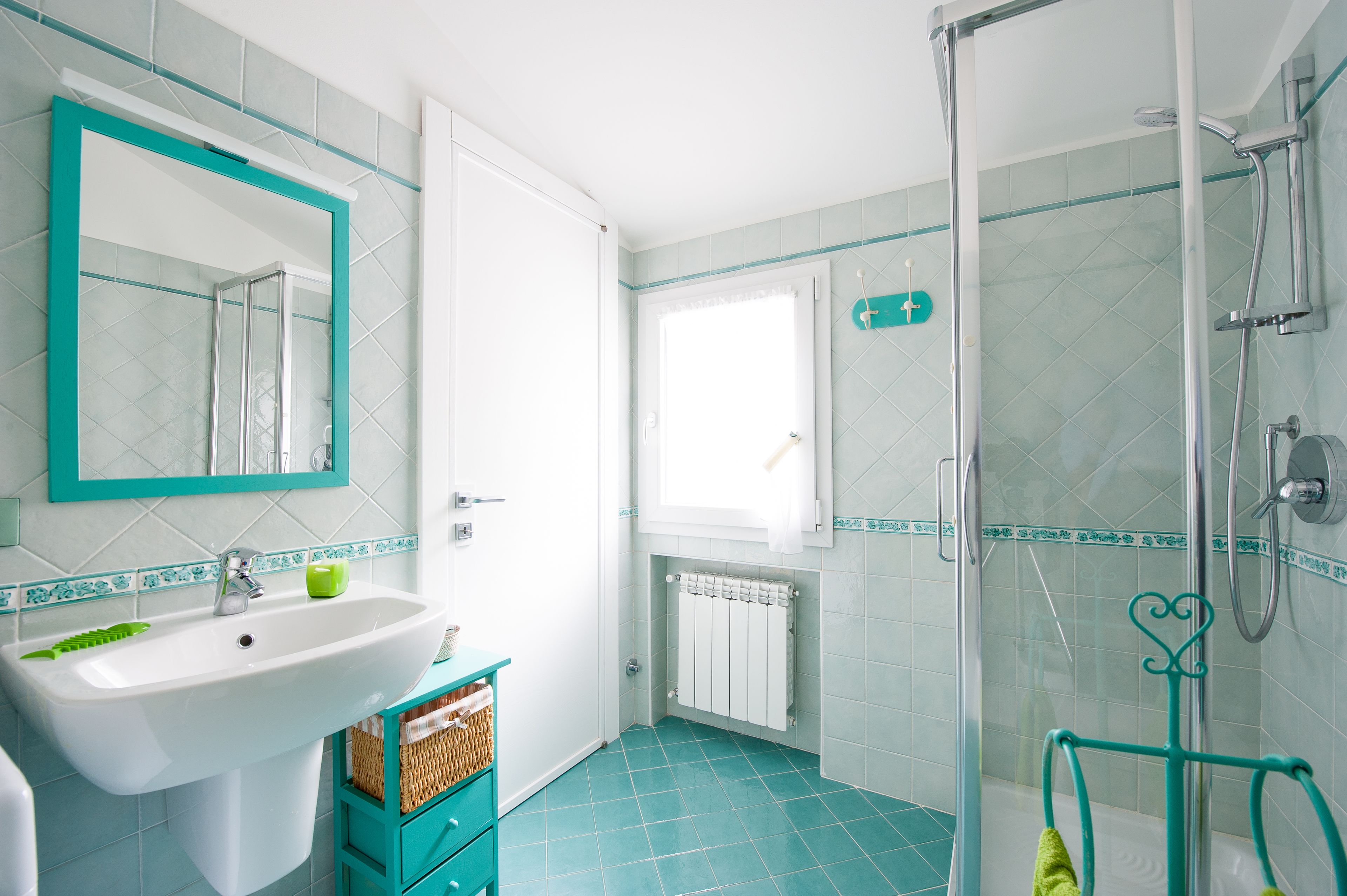 A bathroom with a white sink, a mirror with a teal frame, a shower with a glass door, and a window with a white curtain.