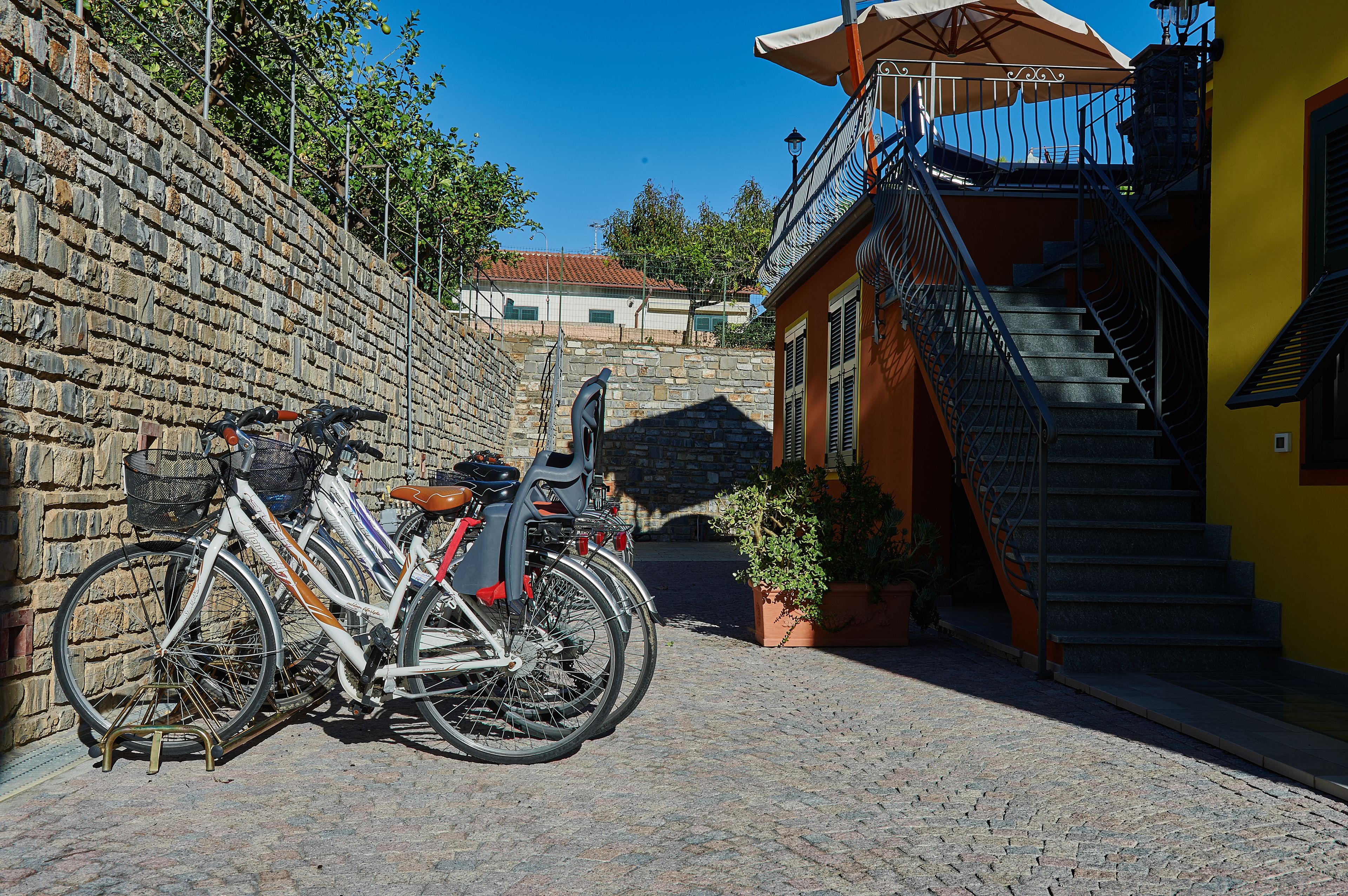 A cobblestone street with bicycles parked against a stone wall, a staircase leading to a balcony, and a building with a yellow facade.