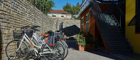 A cobblestone street with bicycles parked against a stone wall, a staircase leading to a balcony, and a building with a yellow facade.