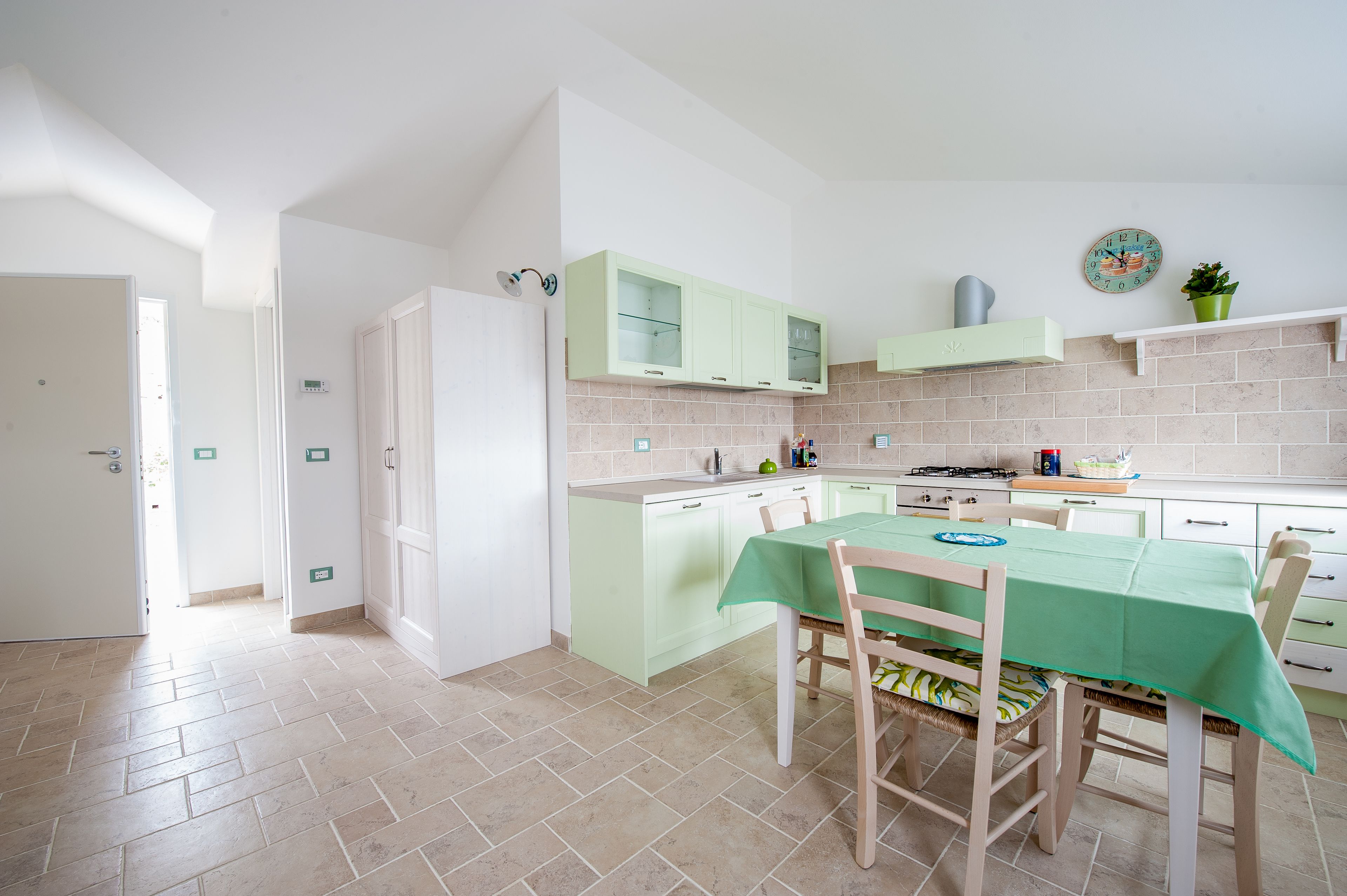 A kitchen with light green cabinets, a dining table with a green tablecloth, and a clock on the wall.