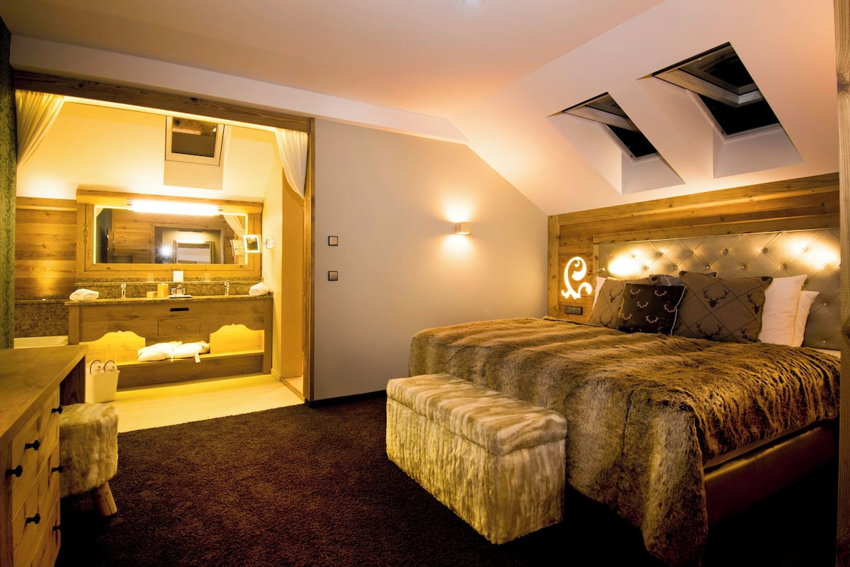 A bedroom with a large bed, a bench, a dresser, and a bathroom with a mirror and sink.