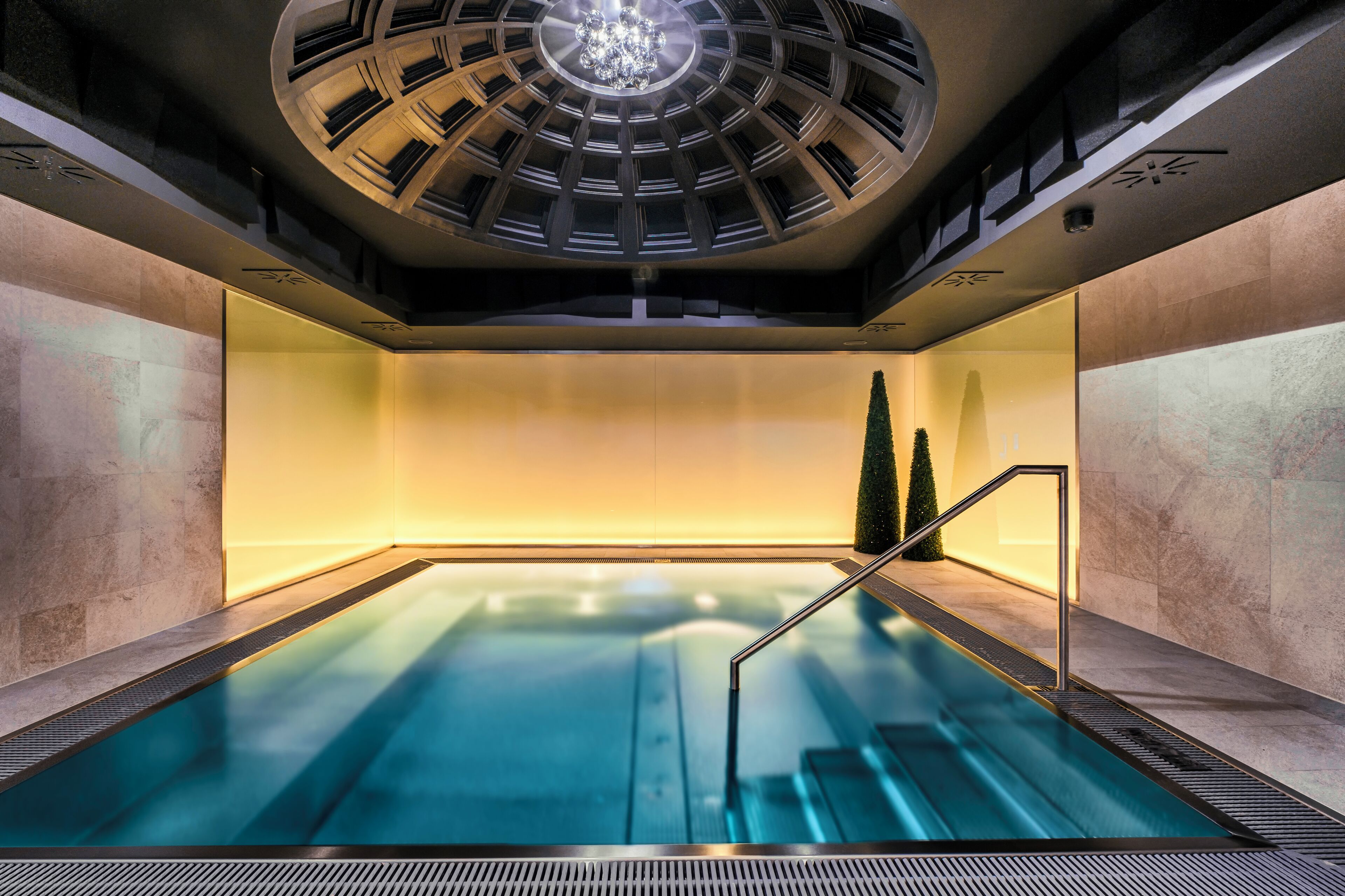 A modern indoor pool with a central water feature, surrounded by stone walls and a skylight above.