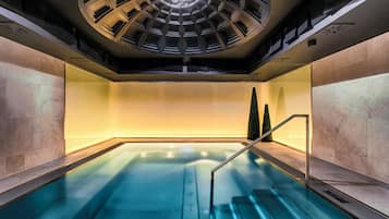 A modern indoor pool with a central water feature, surrounded by stone walls and a skylight above.