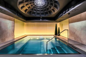 Una piscina interior moderna con un elemento acuático central, rodeada de muros de piedra y un tragaluz.