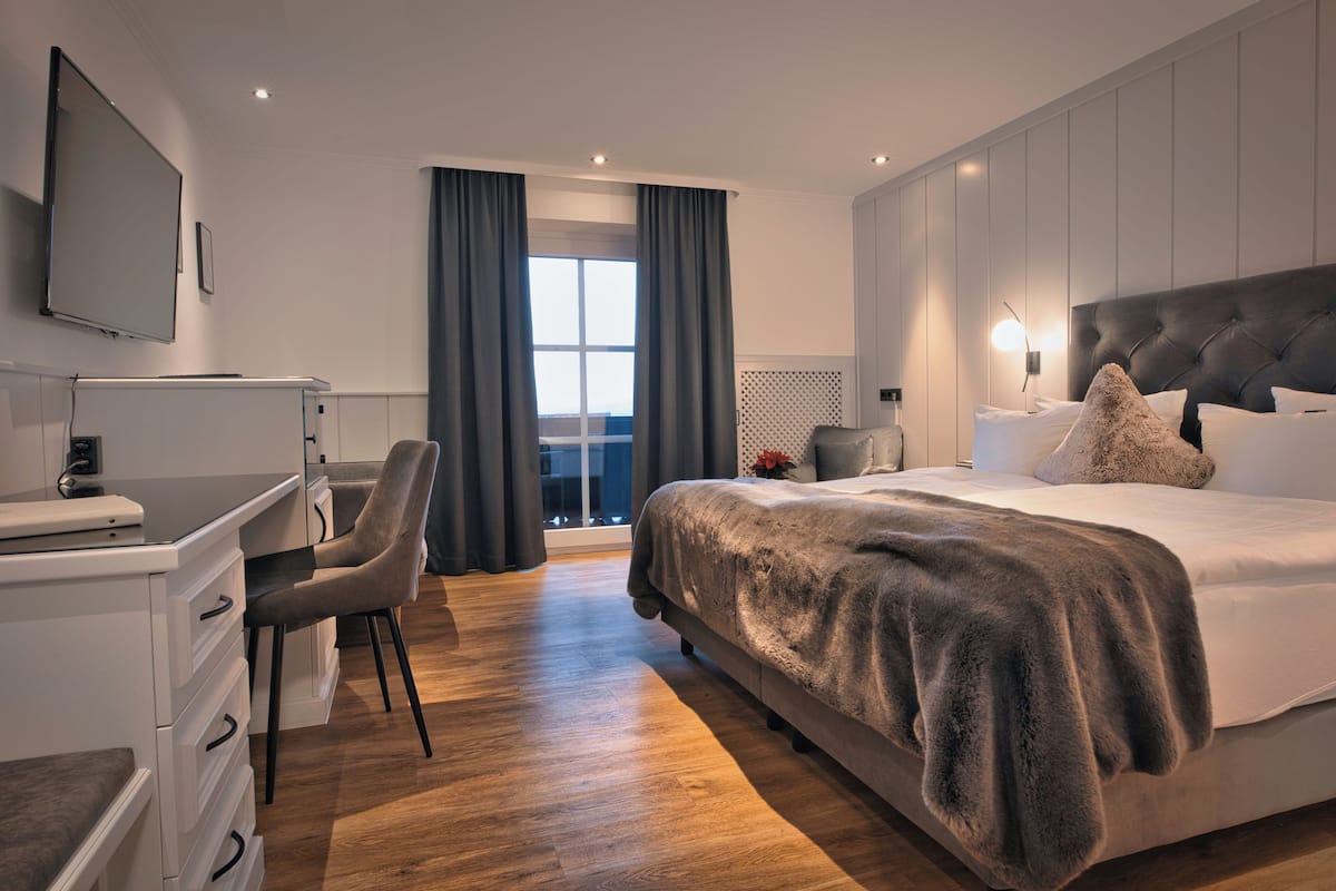 A modern hotel room with a large bed, a desk with a laptop, a chair, and a view of the outdoors.