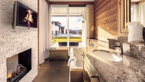 A modern bathroom with a fireplace, a large bathtub, and a view of a building complex. - Landromantik Hotel Oswald Gourmet & SPA (Teisnach)