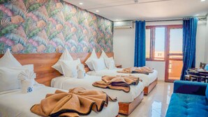 Une chambre d’hôtel avec deux lits, une grande fenêtre avec des rideaux bleus, une tête de lit en bois et un mur décoré d’un motif coloré représentant des feuilles.