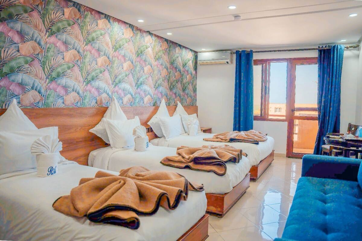 A hotel room with two beds, a large window with blue curtains, a wooden headboard, and a colorful leaf-patterned wall.