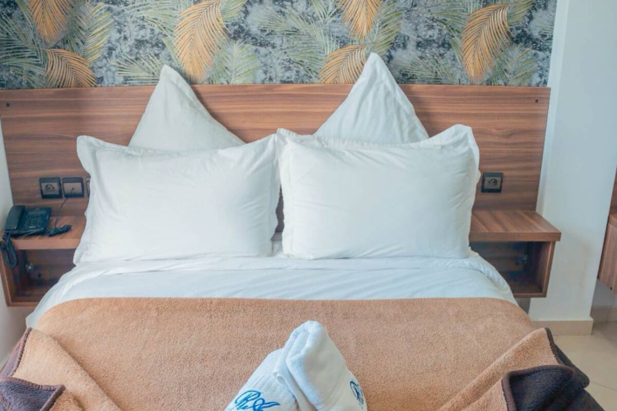 A neatly made bed with a brown blanket and a rolled towel, white pillows, and a wooden headboard.