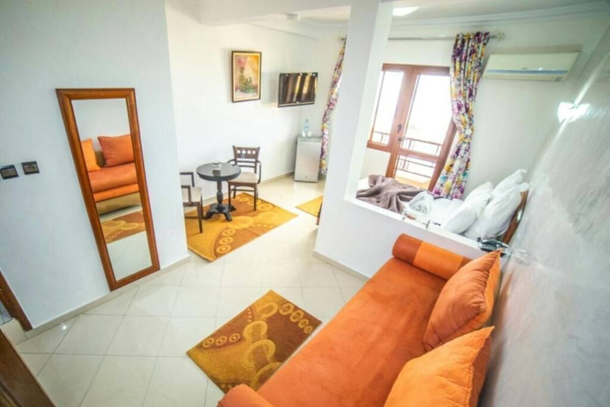 A living room with a sofa, a mirror, a dining table with chairs, a small TV, and a window with curtains.