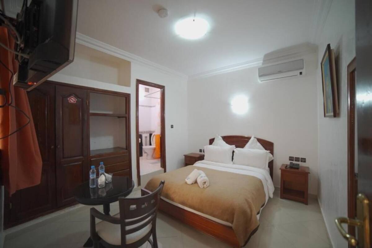 A hotel room with a bed, bedside tables, a desk with chairs, a wardrobe, and an air conditioning unit.