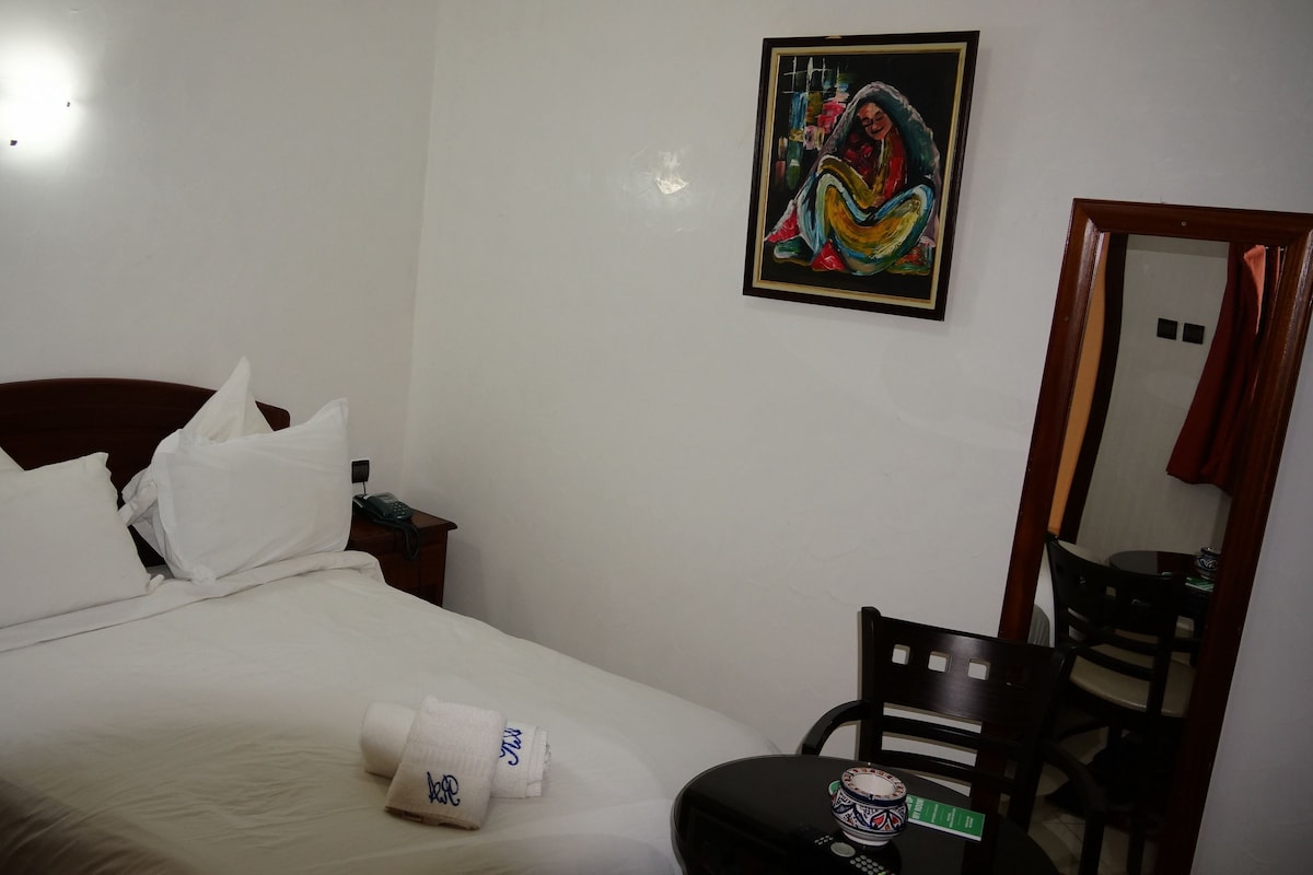 A hotel room with a bed, pillows, a painting on the wall, a chair, and a table with a cup and remote control.