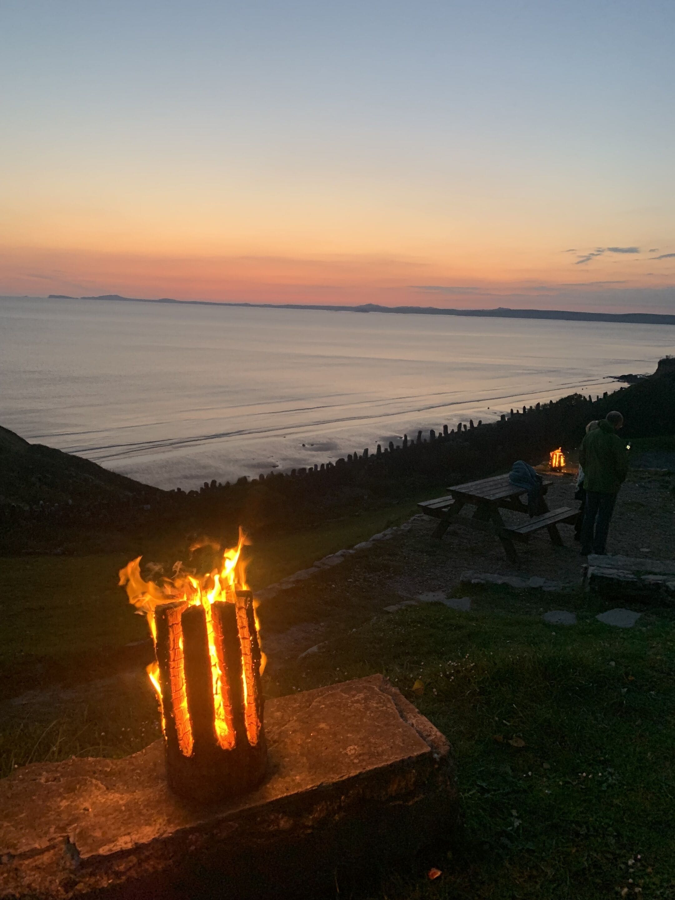 A campfire at dusk with a view of the sea and distant hills.
