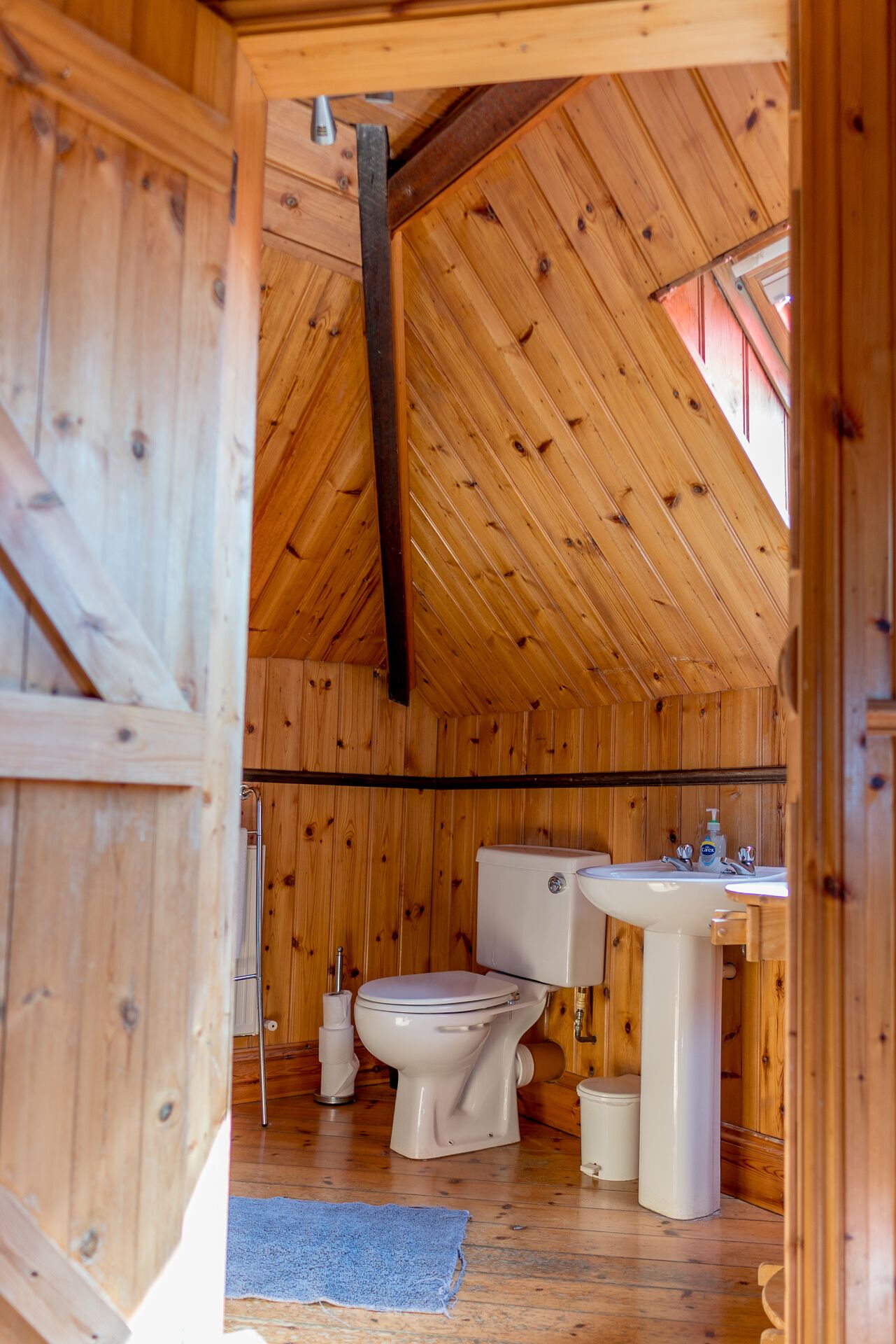 A wooden cabin with a sloped ceiling, a toilet, a sink, and a blue towel on the floor.