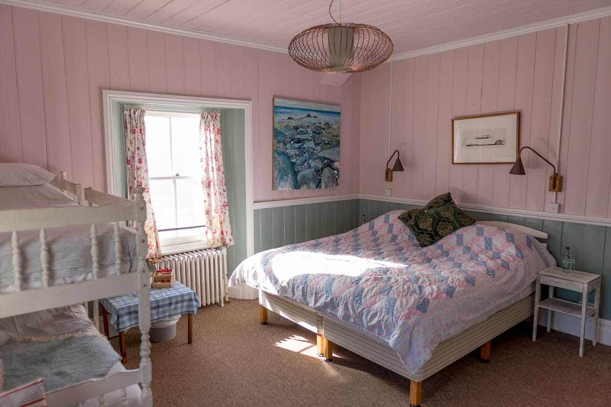 A bedroom with a bunk bed, a single bed, a radiator, a table with a checkered tablecloth, a painting, and a lamp.