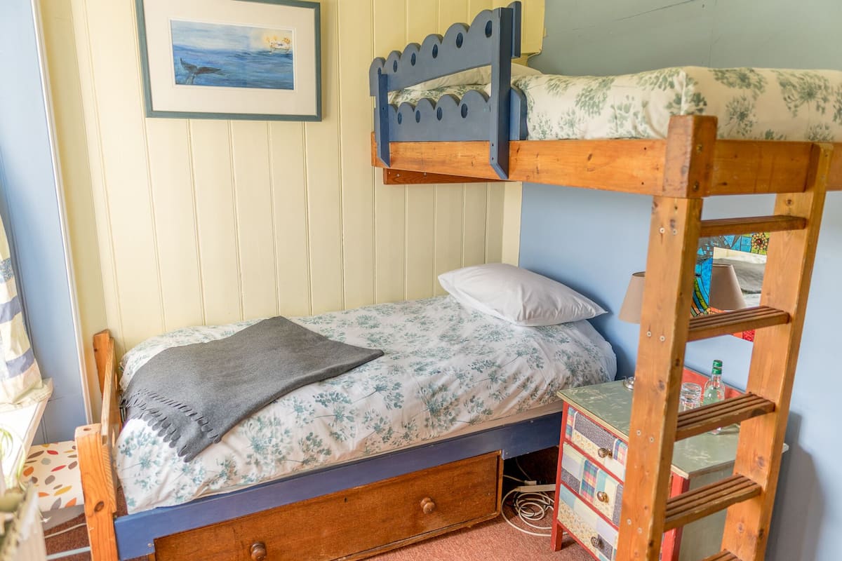 A bunk bed with a wooden ladder, a single bed with floral bedding, a small wooden chest, and a framed picture on the wall.