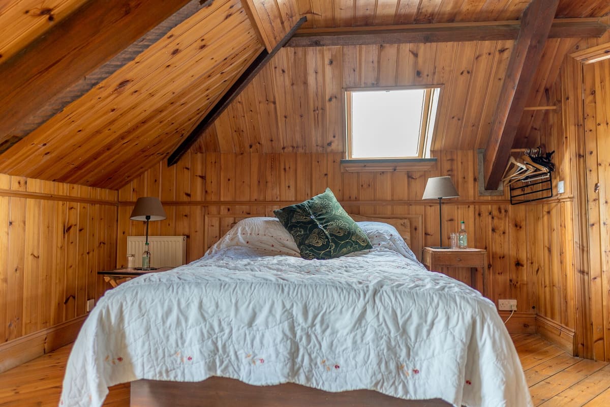 A wooden room with a sloped ceiling, a bed with a white bedspread and a green pillow, two bedside lamps, and a window.