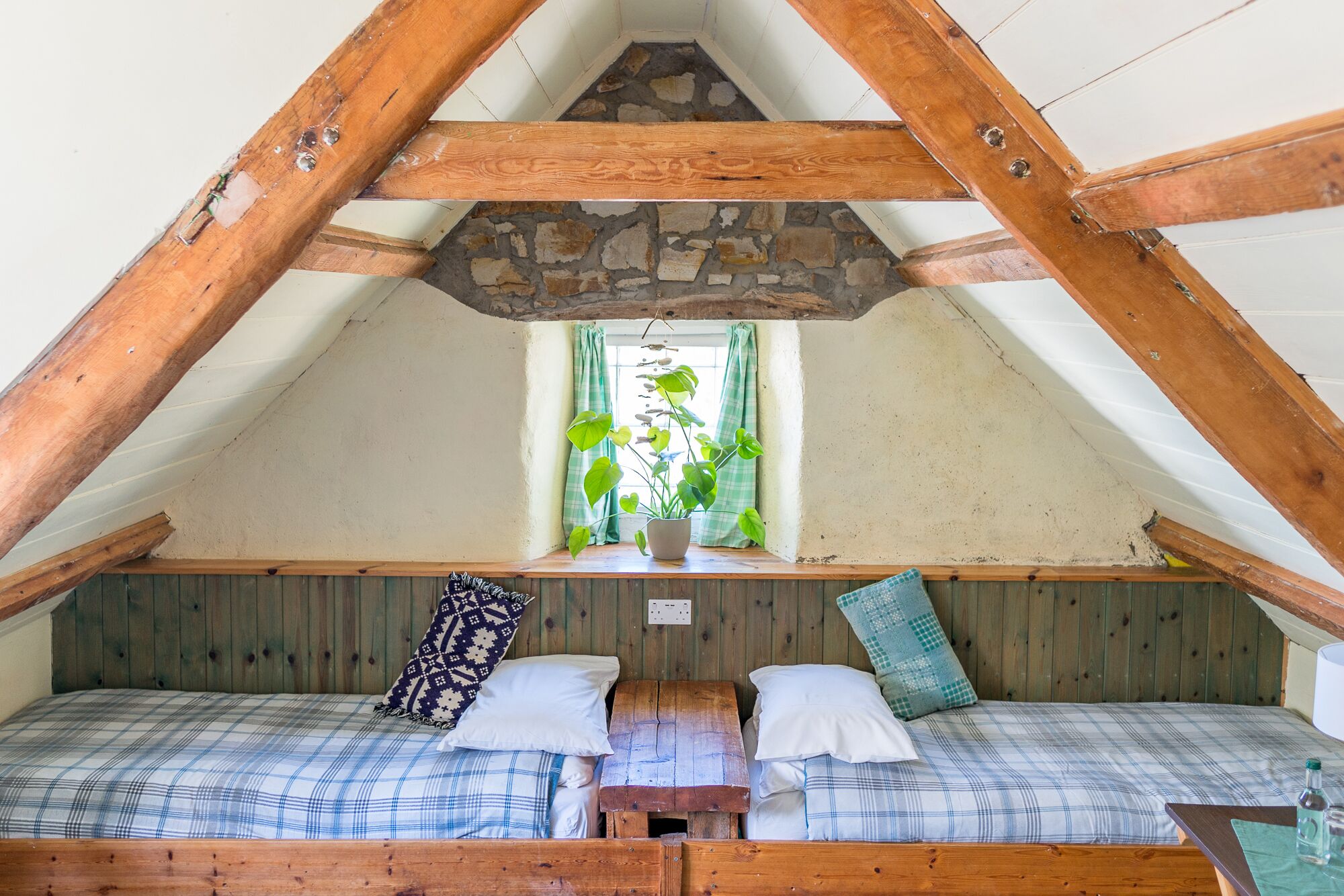 A cozy attic bedroom with a sloped ceiling, wooden beams, a stone wall, and a window with curtains.