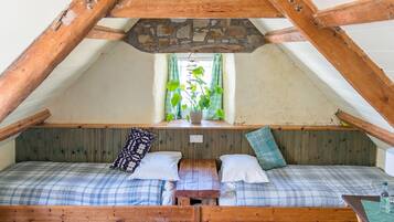 A cozy attic bedroom with a sloped ceiling, wooden beams, a stone wall, and a window with curtains.