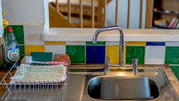 A kitchen with a stainless steel sink, a dish rack, a bottle of Fairy dish soap, and a colorful tiled backsplash.