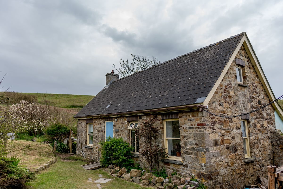 A stone house with a sloped roof, a small porch, and a garden with a wooden fence.