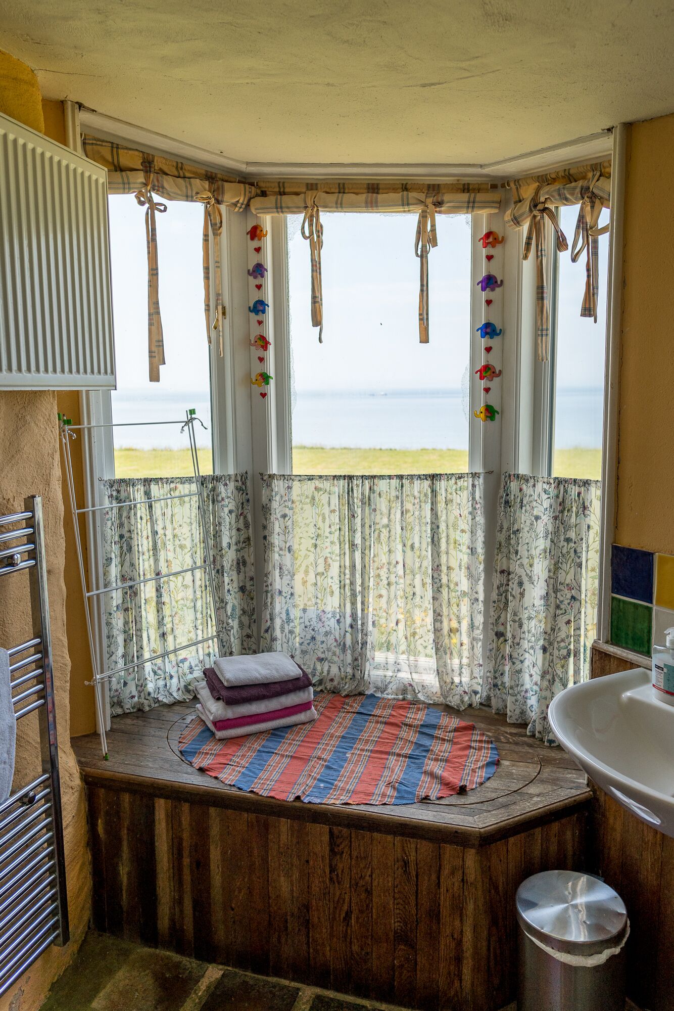 Un bagno piccolo e accogliente con una finestra a bovinda sulla spiaggia. Si notano una panca di legno, un lavandino e un porta asciugamani.