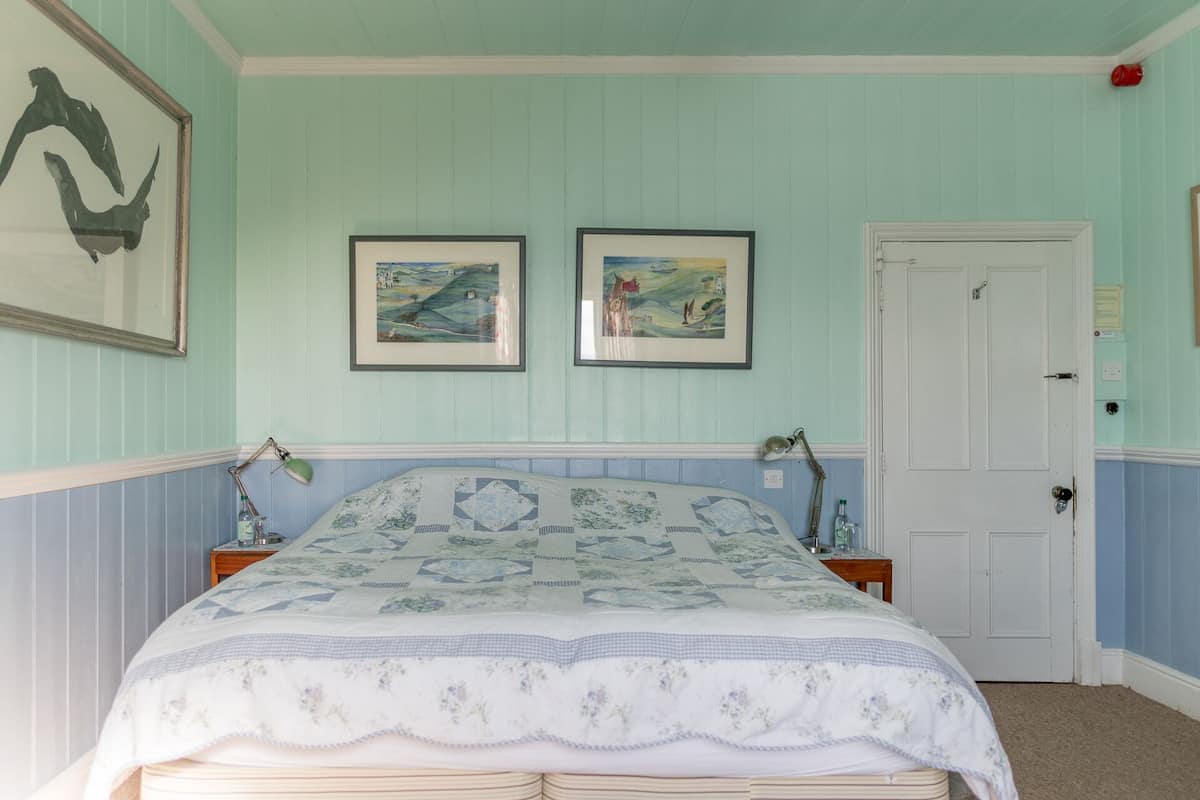 A bedroom with a bed, two framed paintings, a door, and a nightstand with a lamp.