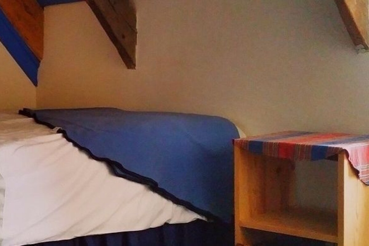 A single bed with a white and blue bedspread, a wooden headboard, and a small wooden table with a colorful tablecloth.
