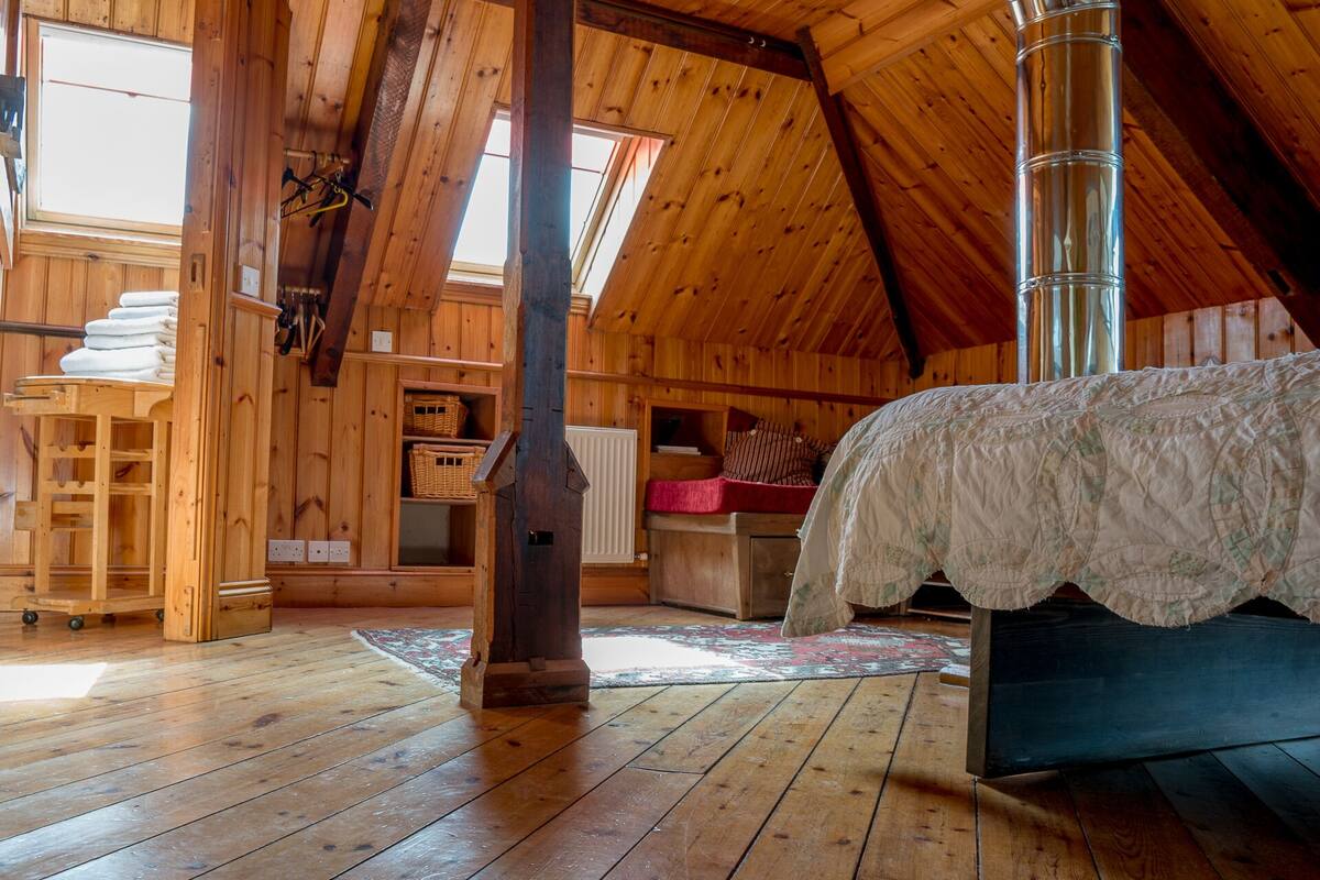 A cozy attic bedroom with wooden walls, a loft bed, a radiator, and a wooden cart.