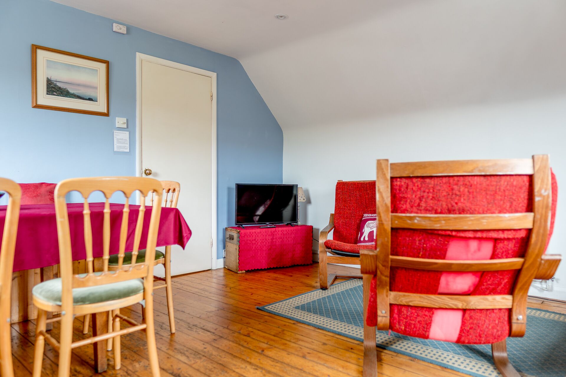 Een kamer met een houten eettafel en stoelen, een televisie op een bijzettafeltje, en een rode fauteuil.