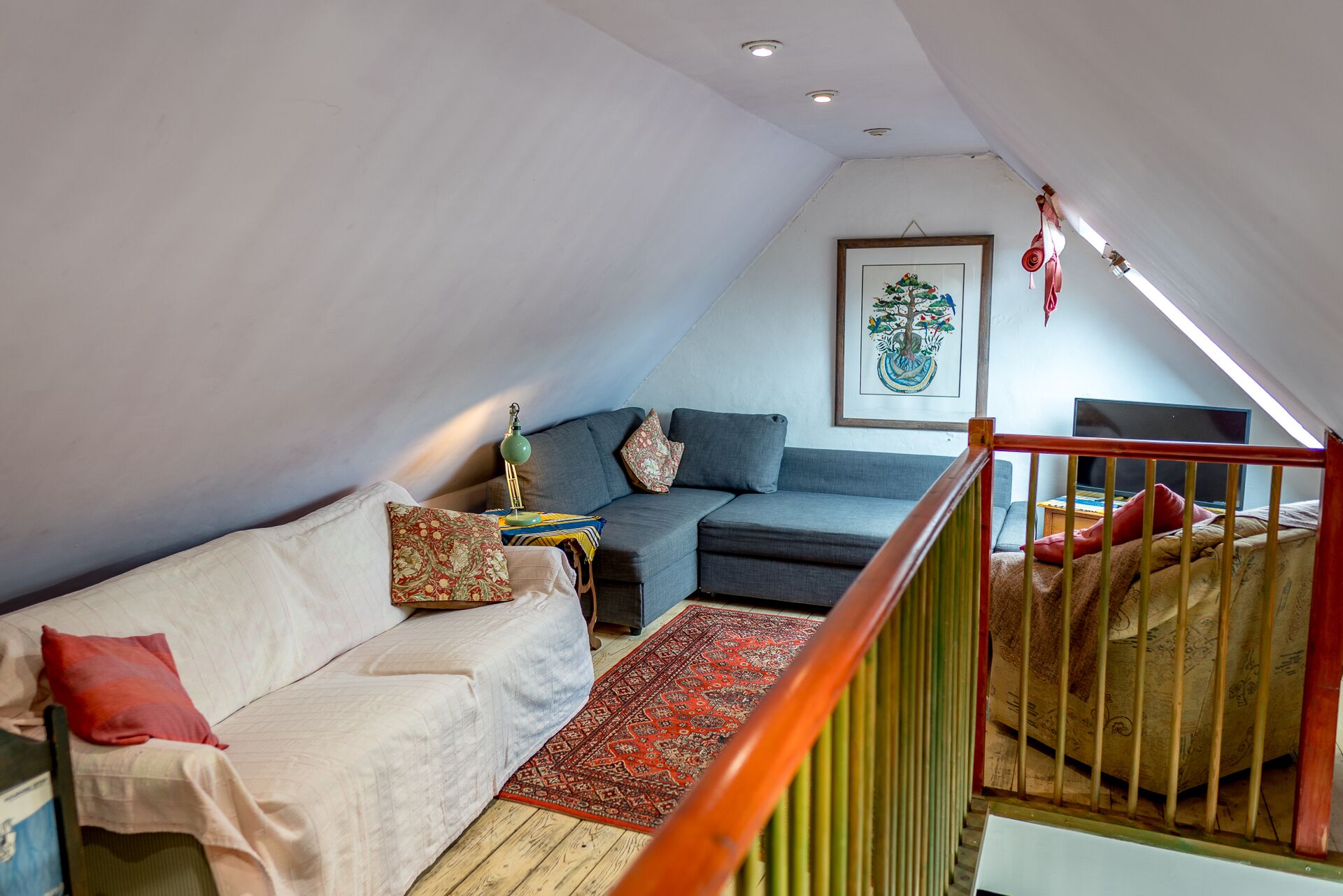 A loft bedroom with a high ceiling, a grey sofa, a white couch, a red patterned rug, and a wooden floor.