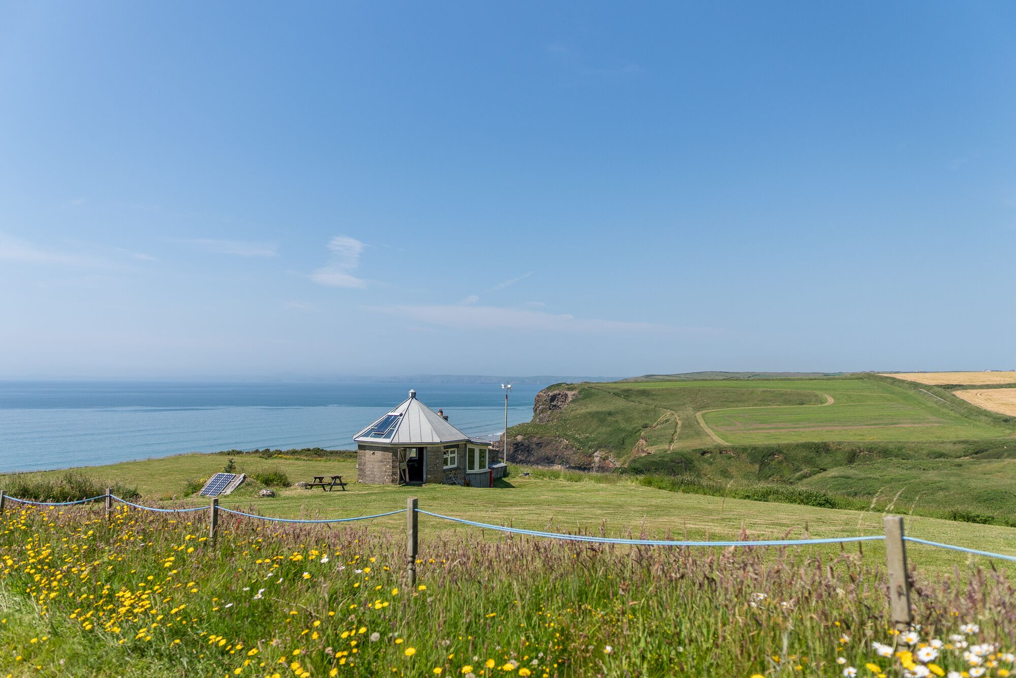 A coastal view with a small stone building, a picnic table, and a fenced area with wildflowers.