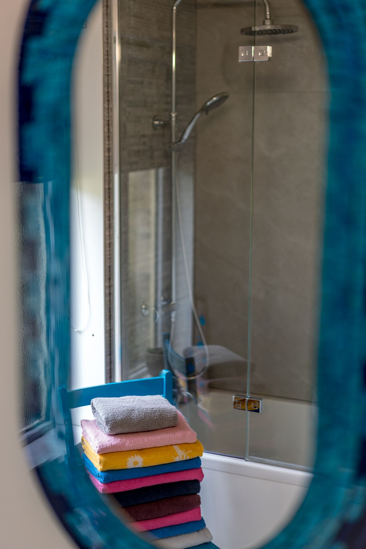A bathroom with a glass shower enclosure, a stack of towels, and a blue chair.