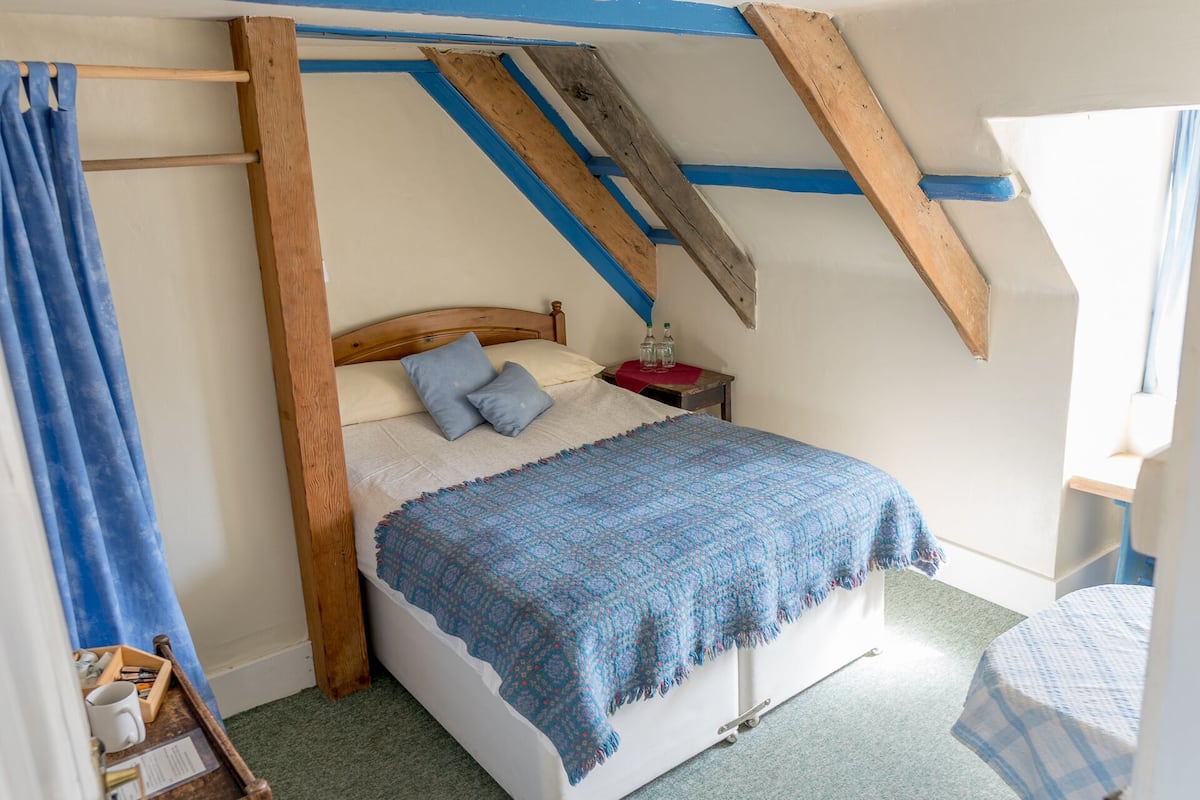 A bedroom with a wooden ceiling, a bed with a blue blanket, a nightstand with bottles, and a window with curtains.