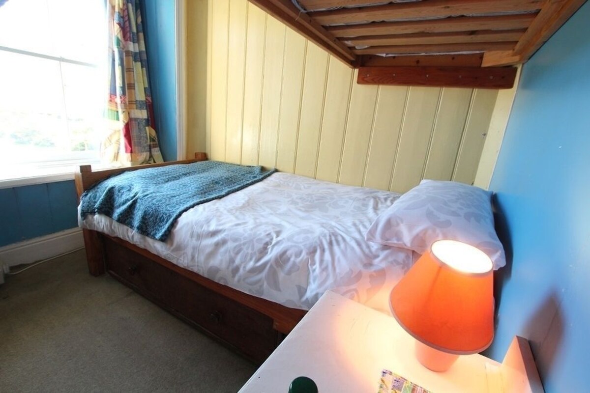 Ein Einzelbett mit Holzrahmen, einer weißen Matratze und einer blauen Decke. Ein Nachttisch mit einer orangefarbenen Lampe steht daneben. Die Wände sind blau und es gibt ein Fenster mit Vorhängen.