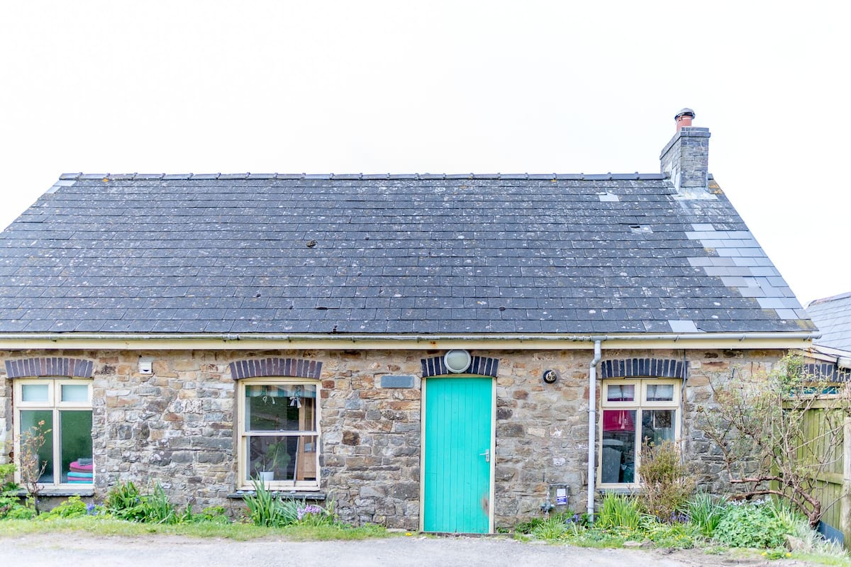 A stone building with a dark slate roof, a turquoise door, and two windows with shutters.