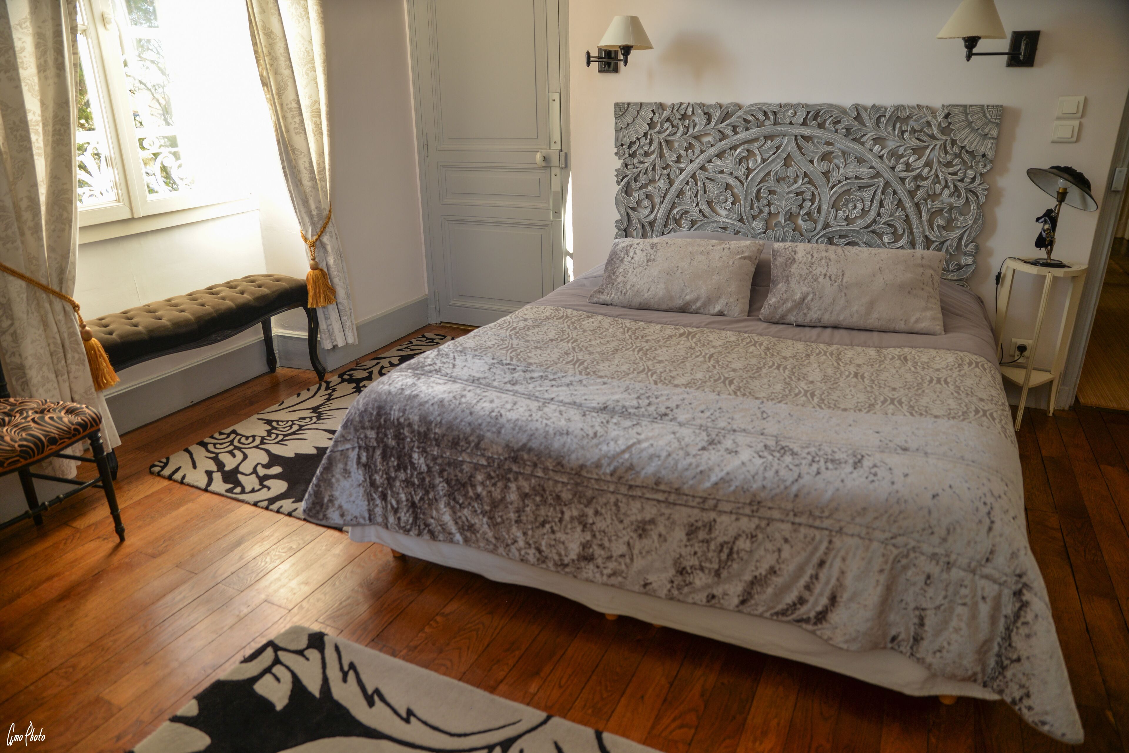 Un dormitorio con una cama grande, un banco, una alfombra estampada y una ventana con cortinas.
