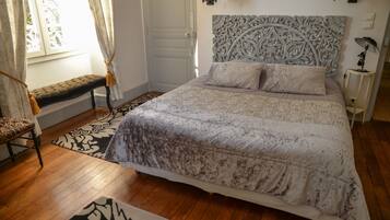 Un dormitorio con una cama grande, un banco, una alfombra estampada y una ventana con cortinas.