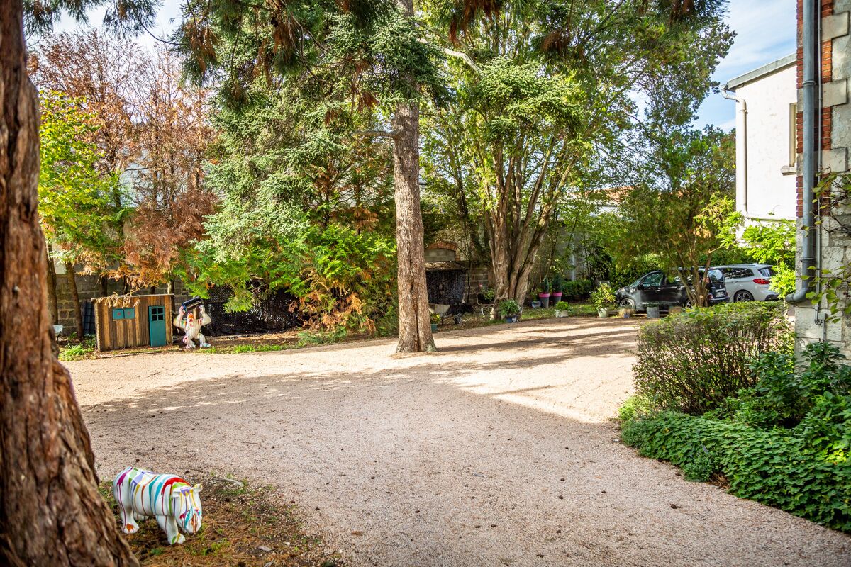 A gravel driveway leading to a building with a white exterior. There are trees and shrubs around, and a small shed in the background.