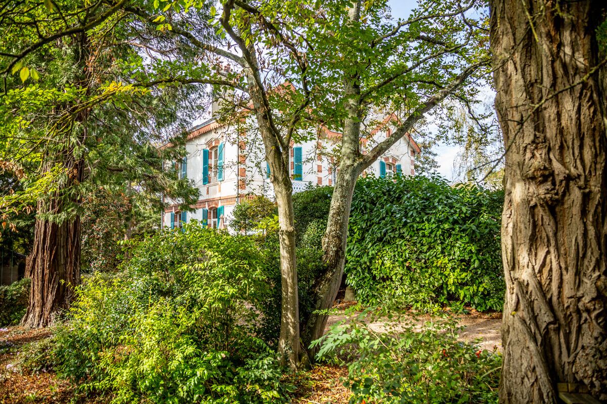 A lush garden with mature trees and a building with red-tiled roof and blue shutters in the background.