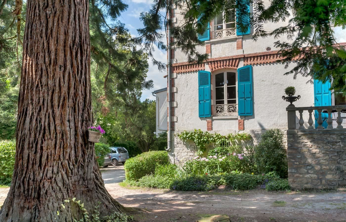 Un edificio rústico con fachada de piedra, contraventanas azules y un gran árbol en primer plano.