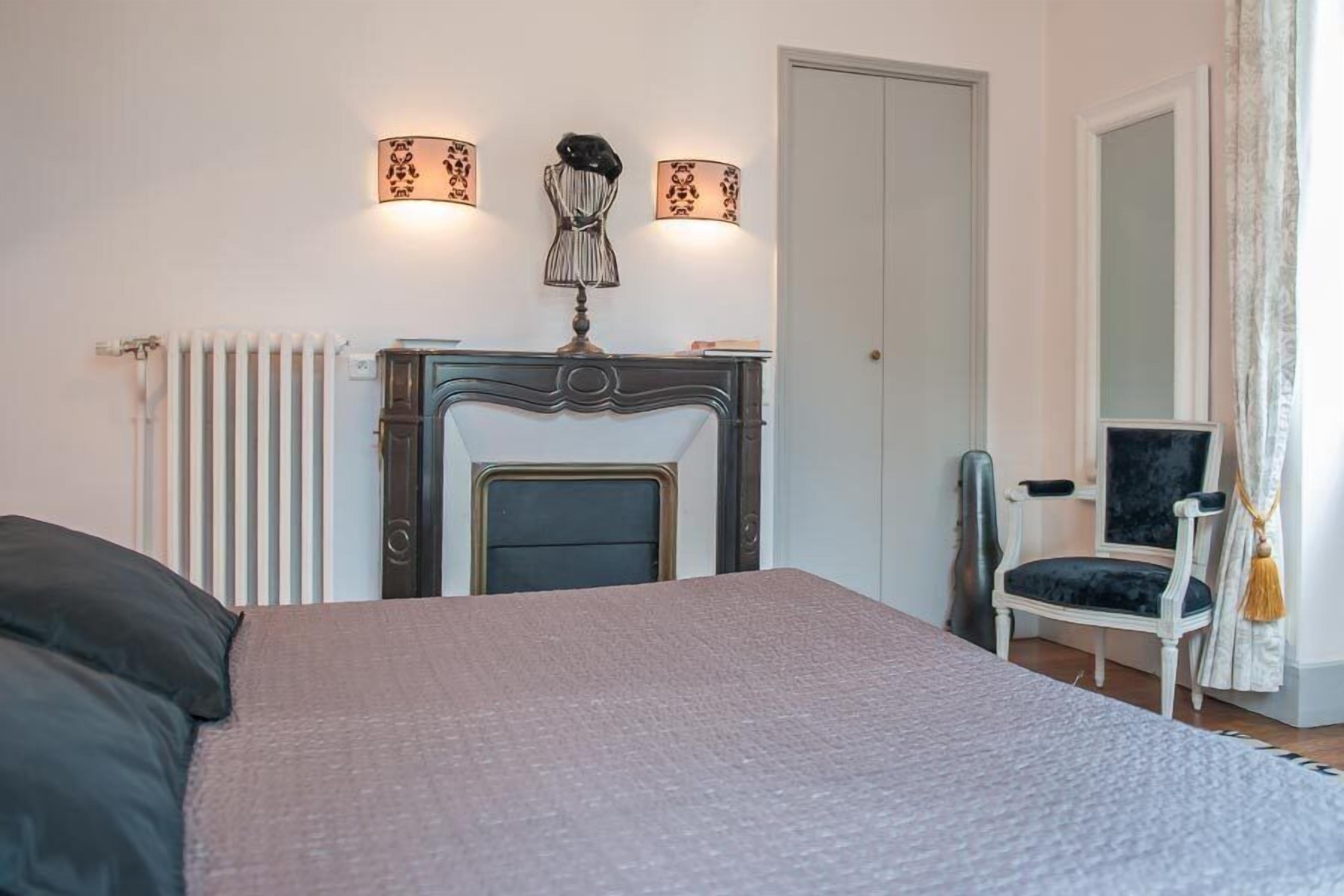 Une chambre à coucher comprenant un lit, une cheminée, un fauteuil et deux lampes fixées au mur.