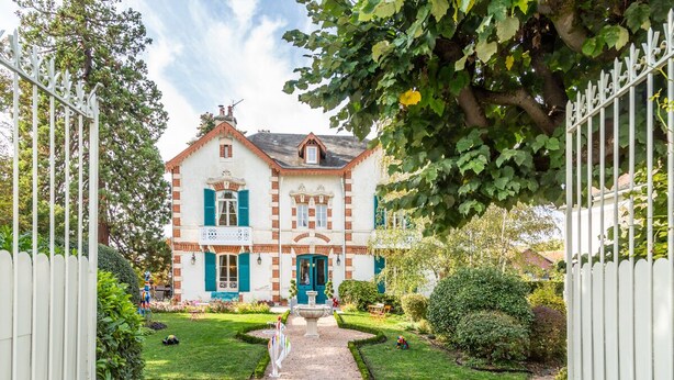 A two-story building with a central entrance, surrounded by a well-maintained garden and a white fence. - L'Oustal (Bourges)