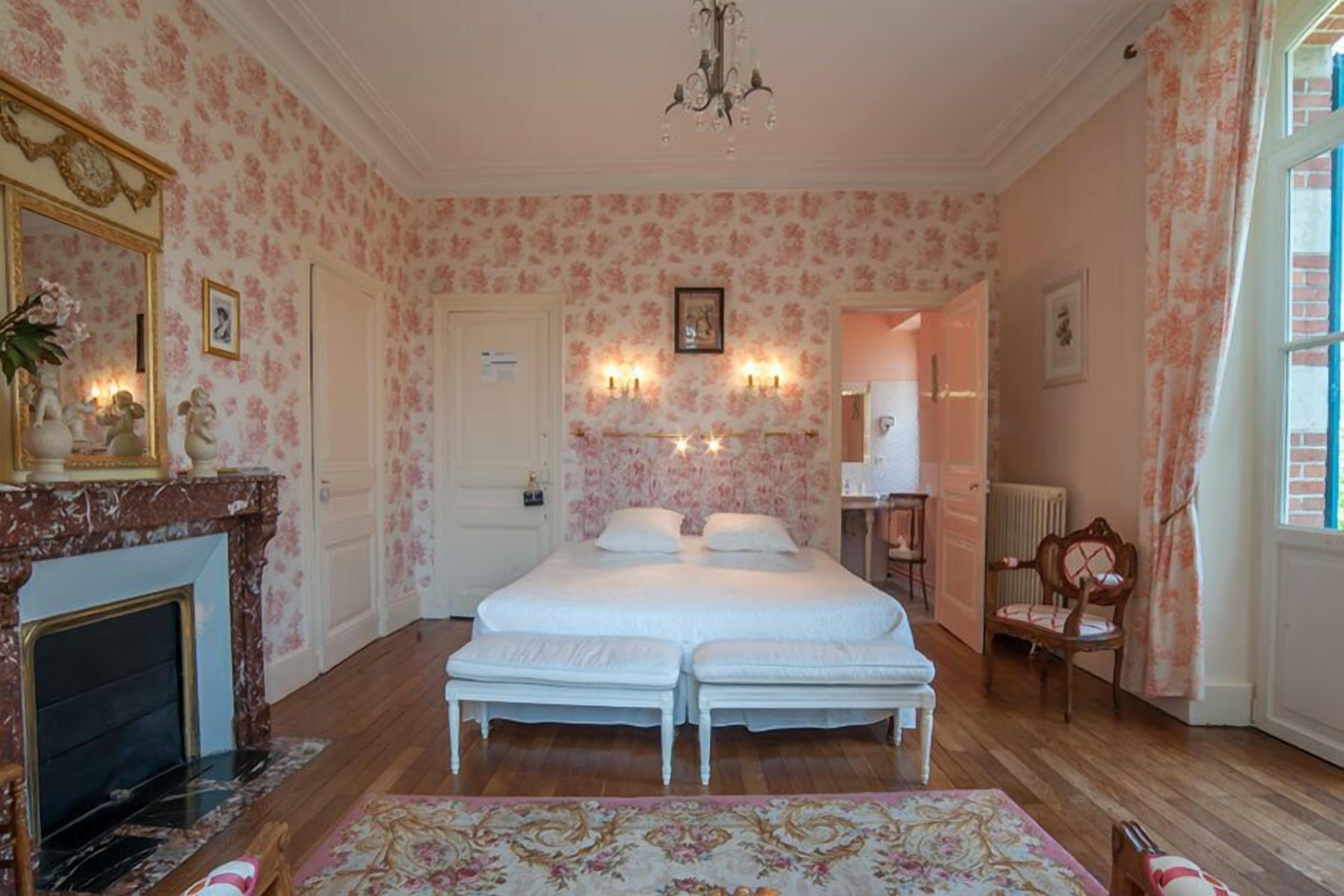 A bedroom with a double bed, a fireplace, a chandelier, and floral wallpaper.