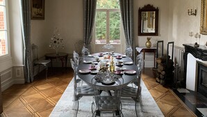 A dining room with a round table set for six, surrounded by clear chairs. A chandelier hangs from the ceiling, and a large window offers a view of greenery outside. - L'Oustal (Bourges)