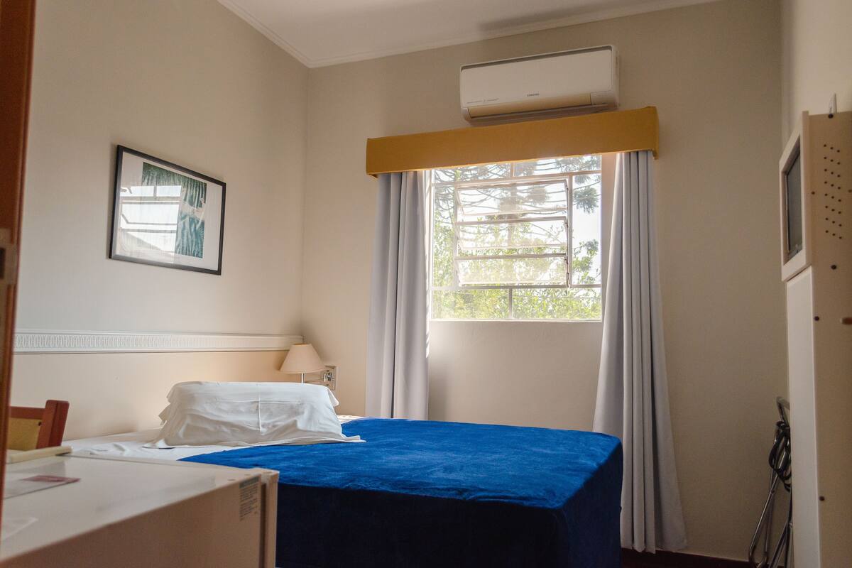 Une chambre d’hôtel comprenant un lit, une table de chevet, une lampe, une fenêtre avec des rideaux et un appareil de climatisation.